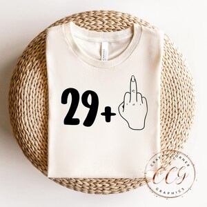 Puede incluir: Una camiseta blanca con texto negro que dice "29+" y un contorno negro de un dedo medio.