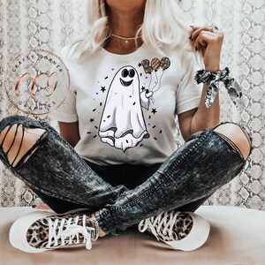 K&ouml;nnte beinhalten: Ein wei&szlig;es T-Shirt mit einem Cartoon-Geist, der Ballons h&auml;lt, auf denen "BOO" steht. Der Geist hat ein l&auml;chelndes Gesicht und ist von Konfetti und Sternen umgeben.