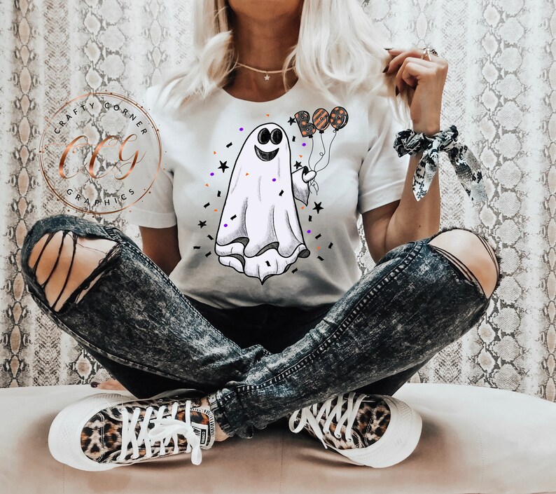 K&ouml;nnte beinhalten: Ein wei&szlig;es T-Shirt mit einem Cartoon-Geist, der Ballons h&auml;lt, auf denen "BOO" steht. Der Geist hat ein l&auml;chelndes Gesicht und ist von Konfetti und Sternen umgeben.