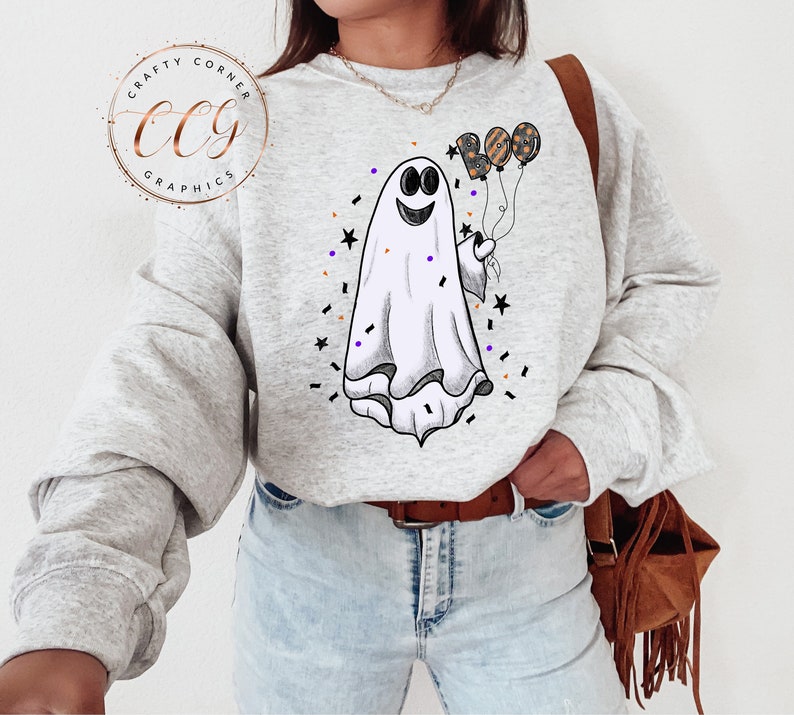 K&ouml;nnte beinhalten: Ein graues Sweatshirt mit einem wei&szlig;en Geist, der drei Ballons h&auml;lt, die "BOO" in Schwarz und Orange buchstabieren. Der Geist hat ein fr&ouml;hliches Gesicht und ist von schwarzen und orangefarbenen Konfetti umgeben.