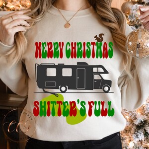 May include: A white sweatshirt with a green, red and white graphic that says "Merry Christmas Shitter's Full". The graphic also includes a black and white illustration of a camper van.