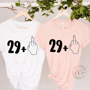 Puede incluir: Dos camisetas con el texto "29+" y un emoji de dedo medio. Las camisetas son blancas y rosas.