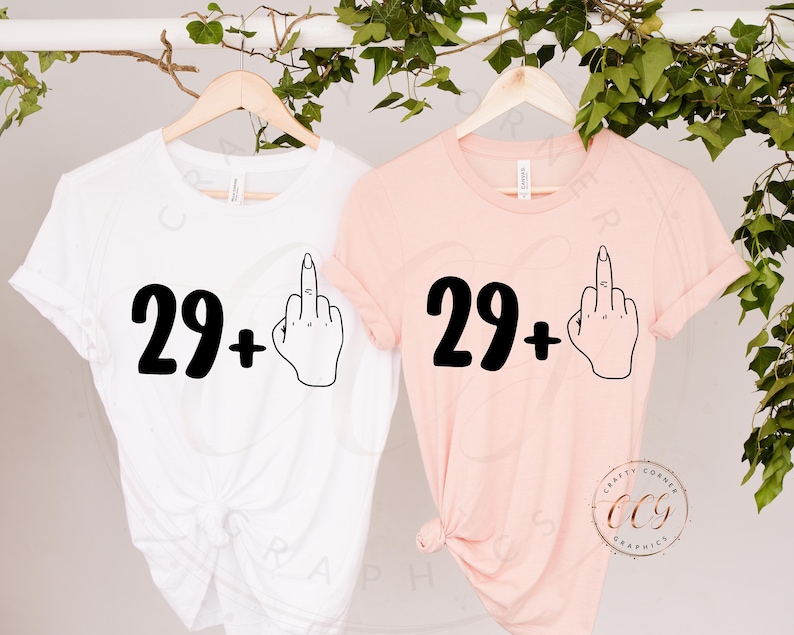 Puede incluir: Dos camisetas con el texto "29+" y un emoji de dedo medio. Las camisetas son blancas y rosas.