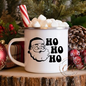 Peut inclure: Mug en &eacute;mail blanc avec une illustration en noir et blanc du P&egrave;re No&euml;l et le texte "HO HO HO". La tasse est remplie de guimauves et d'une canne en sucre.