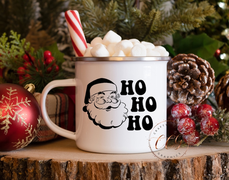 Peut inclure: Mug en &eacute;mail blanc avec une illustration en noir et blanc du P&egrave;re No&euml;l et le texte "HO HO HO". La tasse est remplie de guimauves et d'une canne en sucre.