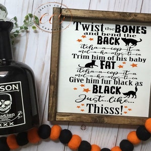 May include: A wooden sign with a black and white Halloween-themed design. The sign reads "Twist the BONES and bend the BACK itcha-a-cop-it-a mel-a-ka-mys-ti-ca Trim him of his baby FAT itcha-a-cop-it-a mel-a-ka-mys-ti-ca Give him fur black as BLACK Just...Like...Thisss!"