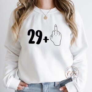 Puede incluir: Sudadera blanca con texto negro que dice "29+" y un dibujo de l&iacute;nea negra de un dedo medio.