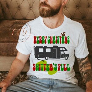 May include: White t-shirt featuring a black and white illustration of a camper van and the text "Merry Christmas Shitter's Full" in red, green, and white.