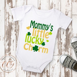May include: White baby onesie with the text "Mommy's little lucky charm" in green and yellow lettering with shamrock designs.