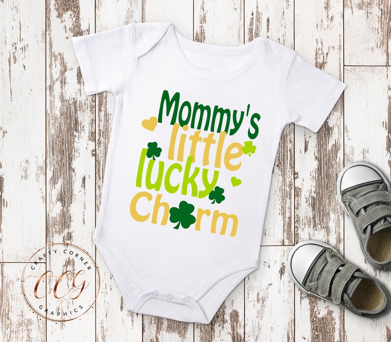 May include: White baby onesie with the text "Mommy's little lucky charm" in green and yellow lettering with shamrock designs.
