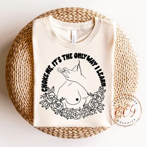 Puede incluir: Camiseta de color crema con un dise&ntilde;o gr&aacute;fico negro. El dise&ntilde;o presenta un dibujo lineal de una mano, senos y una corona floral, con el texto "CHOKE ME. IT'S THE ONLY WAY I LEARN" en disposici&oacute;n circular.