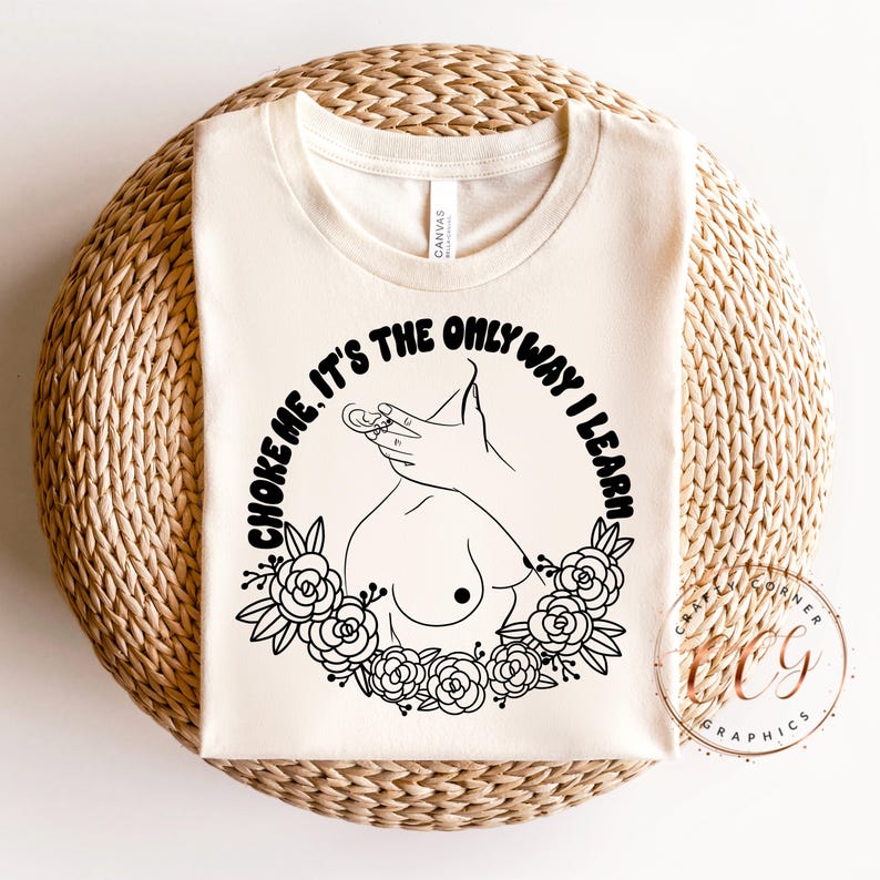 Puede incluir: Camiseta de color crema con un dise&ntilde;o gr&aacute;fico negro. El dise&ntilde;o presenta un dibujo lineal de una mano, senos y una corona floral, con el texto "CHOKE ME. IT'S THE ONLY WAY I LEARN" en disposici&oacute;n circular.