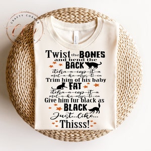 May include: A beige t-shirt with black text that reads "Twist the BONES and bend the BACK itcha-a-cop-it-a mel-a-ka-mys-ti-ca Trim him of his baby FAT itcha-a-cop-it-a mel-a-ka-mys-ti-ca Give him fur black as BLACK Just...Like...Thisss!" with orange stars and black cat silhouettes.