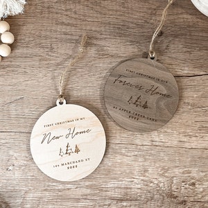 Puede incluir: Dos adornos redondos de madera con un acabado r&uacute;stico. Los adornos est&aacute;n grabados con el texto "Primera Navidad en nuestro hogar para siempre" y "Primera Navidad en mi nuevo hogar", junto con el a&ntilde;o y la direcci&oacute;n.