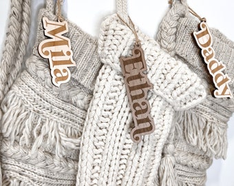 May include: Three knitted stockings with wooden name tags. The tags read "Mila", "Ethan", and "Daddy".