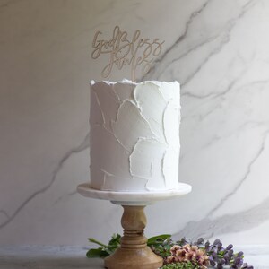 May include: White cake with buttercream frosting on a cake stand. The cake is topped with a wooden sign that says "God Bless Family".