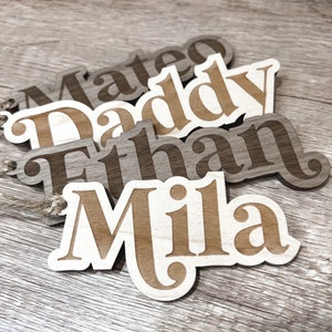 Peut inclure: Quatre &eacute;tiquettes de nom en bois avec les noms Mateo, Daddy, Ethan et Mila. Les &eacute;tiquettes sont brunes et ont un grain de bois brun clair. Les noms sont d&eacute;coup&eacute;s dans le bois et ont un contour blanc.