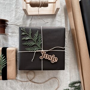 Peut inclure: Une bo&icirc;te cadeau noire avec une brindille de verdure et une &eacute;tiquette cadeau en bois qui dit "Josie". La bo&icirc;te est attach&eacute;e avec de la ficelle et repose sur une surface en lin blanc.