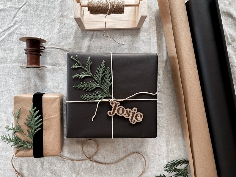 Peut inclure: Une bo&icirc;te cadeau noire avec une brindille de verdure et une &eacute;tiquette cadeau en bois qui dit "Josie". La bo&icirc;te est attach&eacute;e avec de la ficelle et repose sur une surface en lin blanc.