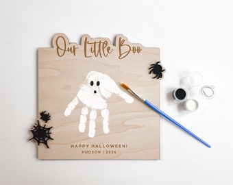 May include: A wooden sign with the text "Our Little Boo" and "Happy Halloween! Hudson | 2024". A white handprint is painted on the sign to look like a ghost. The sign is accompanied by a blue paintbrush, black paint, white paint, and black spider and spiderweb cutouts.