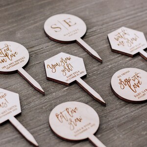May include: Set of 7 wooden cake toppers with various sayings and dates. The toppers are round and hexagonal in shape and feature a brown wood finish. The text on the toppers includes "Rooted in love", "Plant one on us", "You grow girl", "Let love grow", "Watch me grow", "All things grow with love", and "SE".