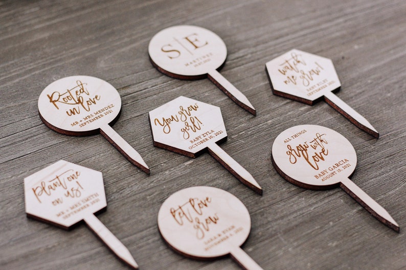 May include: Set of 7 wooden cake toppers with various sayings and dates. The toppers are round and hexagonal in shape and feature a brown wood finish. The text on the toppers includes "Rooted in love", "Plant one on us", "You grow girl", "Let love grow", "Watch me grow", "All things grow with love", and "SE".