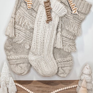 Peut inclure: Trois chaussettes en tricot de couleur cr&egrave;me avec des &eacute;tiquettes en bois portant les noms "Mila", "Ethan" et "Daddy". Les chaussettes sont accroch&eacute;es &agrave; un mur blanc avec une guirlande de perles en bois et de petits arbres blancs sur un sol en bois.