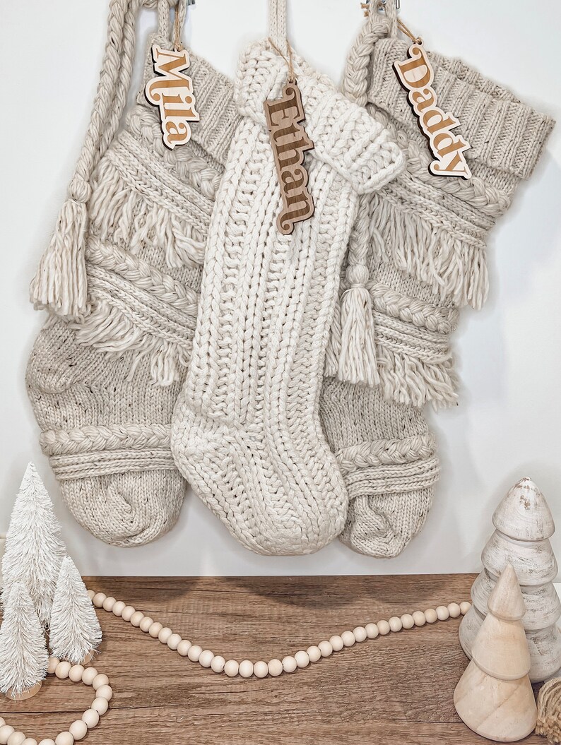 Peut inclure: Trois chaussettes en tricot de couleur cr&egrave;me avec des &eacute;tiquettes en bois portant les noms "Mila", "Ethan" et "Daddy". Les chaussettes sont accroch&eacute;es &agrave; un mur blanc avec une guirlande de perles en bois et de petits arbres blancs sur un sol en bois.
