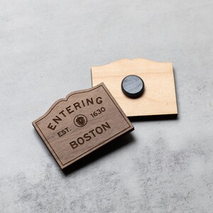 May include: Wooden sign with the text "Entering Est. 1630 Boston" with a small graphic of a tree. The sign is attached to a wooden backing with a magnet.