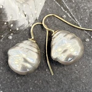 Op de afbeelding: Een paar elegante oorhangers met grote, onregelmatig gevormde, glanzende witte parels. De parels hangen aan delicate goudkleurige oorhaken, wat een verfijnd en tijdloos sieraad creëert.
