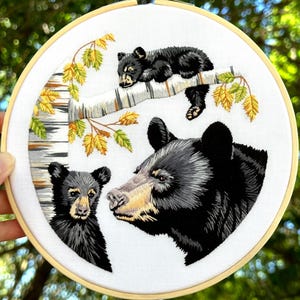 May include: Embroidered hoop art featuring a black bear and two cubs. The bear is facing the viewer with its head tilted slightly to the side. The cubs are nestled in a tree branch with leaves in shades of green and yellow. The background is white.