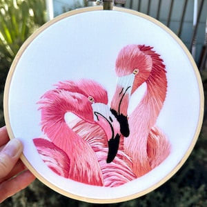 May include: Embroidered hoop art featuring two pink flamingos with their heads intertwined. The flamingos are depicted in a realistic style with detailed feathers and black beaks.