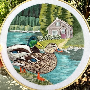 May include: Embroidered artwork of a lake scene with two ducks. The ducks are brown and green with orange beaks and feet. The lake is blue and green with a small house in the background.