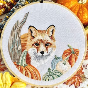 May include: Embroidered hoop art featuring a fox with orange and white fur, surrounded by embroidered pumpkins and gourds in shades of orange, green, and brown.