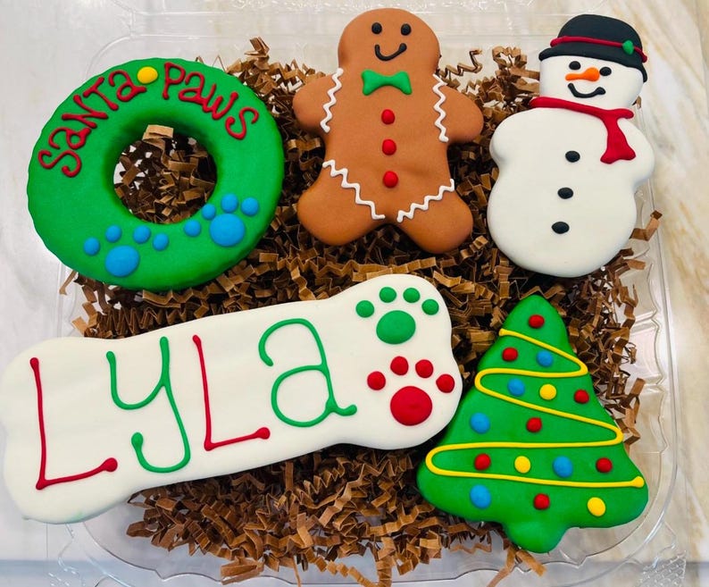 Puede incluir: Una colecci&oacute;n de golosinas para perros decoradas en un recipiente de pl&aacute;stico transparente. Las golosinas incluyen una corona verde con "Santa Paws", un hombre de jengibre, un mu&ntilde;eco de nieve, un hueso con "LYLA" y un &aacute;rbol de Navidad.
