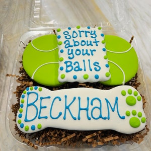 May include: A white bone-shaped cookie decorated with blue and green icing and the text "Beckham". A green tennis ball-shaped cookie with white trim is decorated with blue and green icing and the text "Sorry about your balls".
