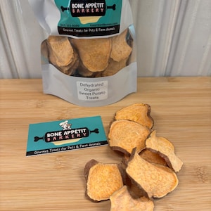 May include: A bag of dehydrated organic sweet potato treats for pets and farm animals. The bag is white with a brown and white dog illustration and the text "Bone Appetit Barkery". The bag is open and there are several sweet potato treats on a wooden surface.