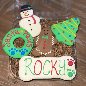 Puede incluir: Una caja transparente llena de golosinas para perros. Las golosinas incluyen un mu&ntilde;eco de nieve, un hombre de jengibre, un &aacute;rbol de Navidad, un hueso con el nombre "Rocky" y una corona verde con "Santa Paws".