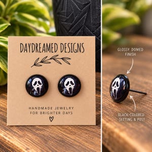 May include: Round stud earrings featuring a white ghost mask graphic on a dark background. The earrings have a glossy domed finish and a black-colored setting and post. They are displayed on a tan card labeled Daydreamed Designs.