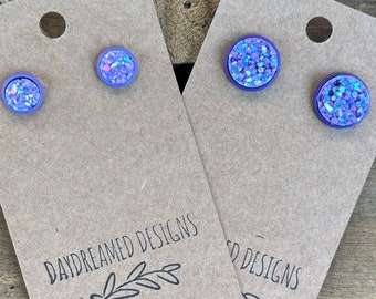 May include: Two pairs of round stud earrings featuring a purple bezel setting and a textured, iridescent, light-reflecting surface. The earrings are mounted on two light brown rectangular display cards with the text DAYDREAMED DESIGNS printed.