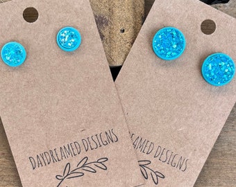 May include: Two pairs of circular stud earrings featuring a bright turquoise resin base with a textured, shimmering blue and silver glitter inlay. The earrings are mounted on brown rectangular display cards labeled Daydreamed Designs.
