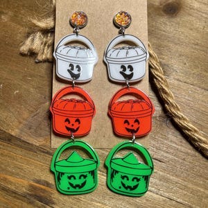 Op de afbeelding: Hangende oorbellen met drie gestapelde emmervormige bedels in wit, oranje en groen. Elke bedel heeft een uniek gezichtje. De bovenkant is een ronde oorsteker met oranje glitters. De bedels zijn verbonden door zilverkleurige metalen ringetjes.