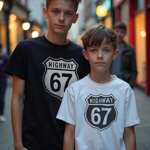 May include: Two people wearing black and white t-shirts featuring a Highway 67 graphic. The graphic is a black and white shield with the word "HIGHWAY" above the number "67". They are standing on a street with shops in the background.