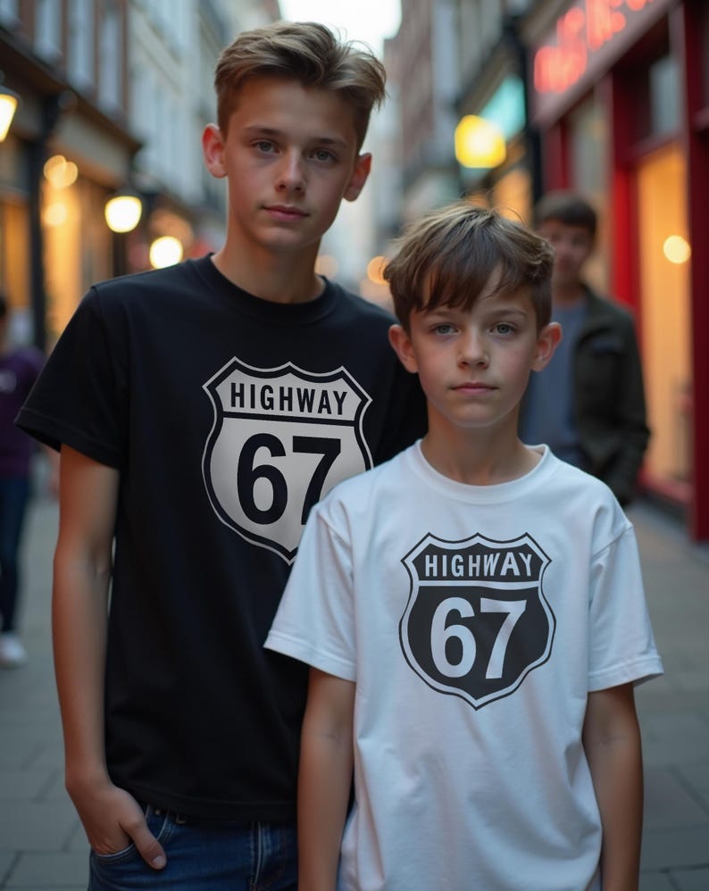 May include: Two people wearing black and white t-shirts featuring a Highway 67 graphic. The graphic is a black and white shield with the word "HIGHWAY" above the number "67". They are standing on a street with shops in the background.