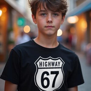 May include: A person wearing a black t-shirt featuring a white graphic of a highway sign with the text "HIGHWAY 67". The person has brown hair and is looking at the camera. The background is blurred, with warm tones.