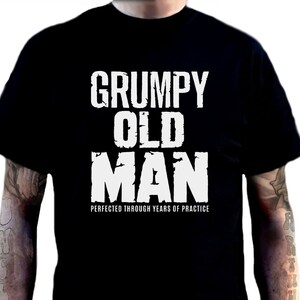 Pode incluir: Camiseta preta com o texto "GRUMPY OLD MAN" em letras brancas grandes. Abaixo, a frase "PERFECTED THROUGH YEARS OF PRACTICE" em letras brancas menores. Camisetas adicionais em várias cores são exibidas à direita.