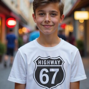 May include: A white t-shirt with a black and white Highway 67 graphic. The graphic is a shield shape with the word "HIGHWAY" above the number "67". The person is smiling.