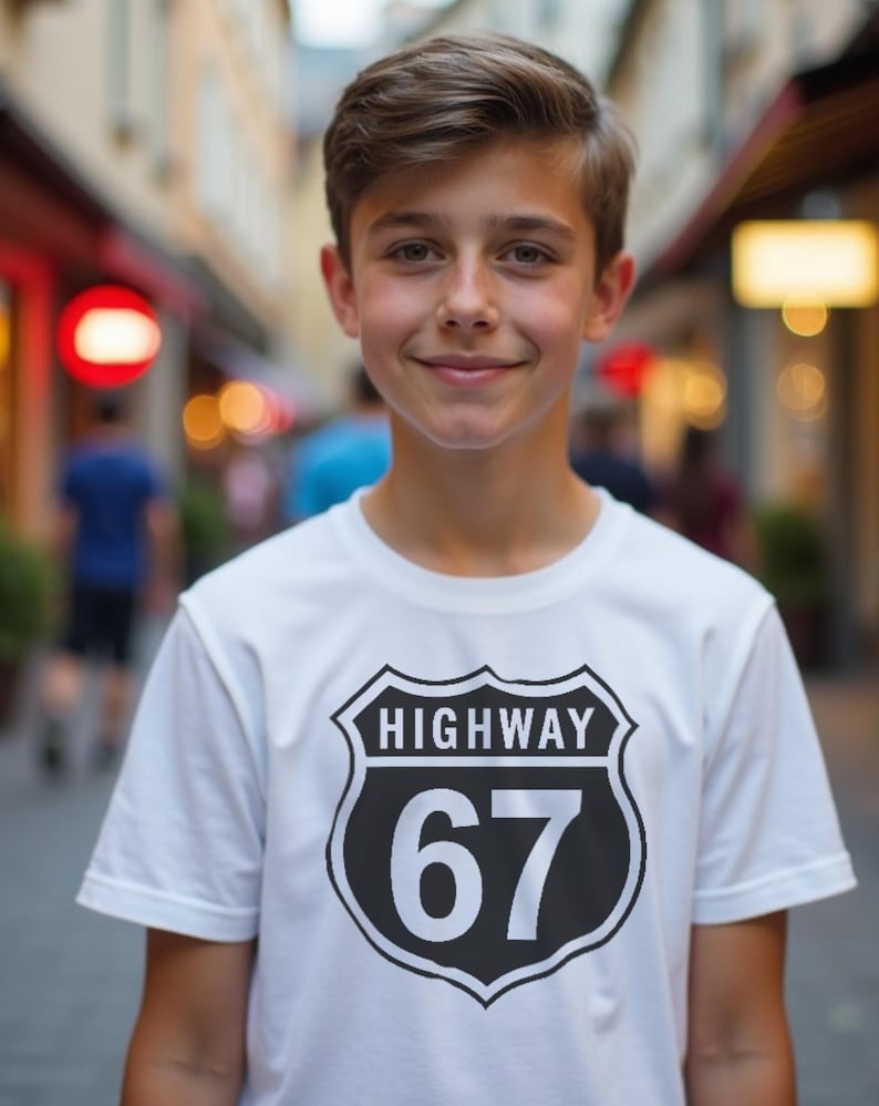 May include: A white t-shirt with a black and white Highway 67 graphic. The graphic is a shield shape with the word "HIGHWAY" above the number "67". The person is smiling.