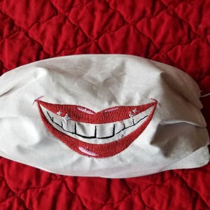 Puede incluir: Máscara facial blanca con una sonrisa bordada en rojo. La sonrisa tiene dientes blancos y un contorno rosa.