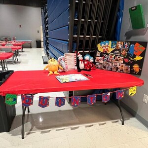 Pu&ograve; includere: Un tavolo rosso con una tovaglia rossa e un libro sopra. C'&egrave; uno striscione che dice "QUINTIN" sul tavolo. Ci sono anche animali di peluche sul tavolo, tra cui il Gatto con il Cappello, Thing 1, Thing 2 e il Lorax. C'&egrave; un cartello che dice "We Love You Quintin".